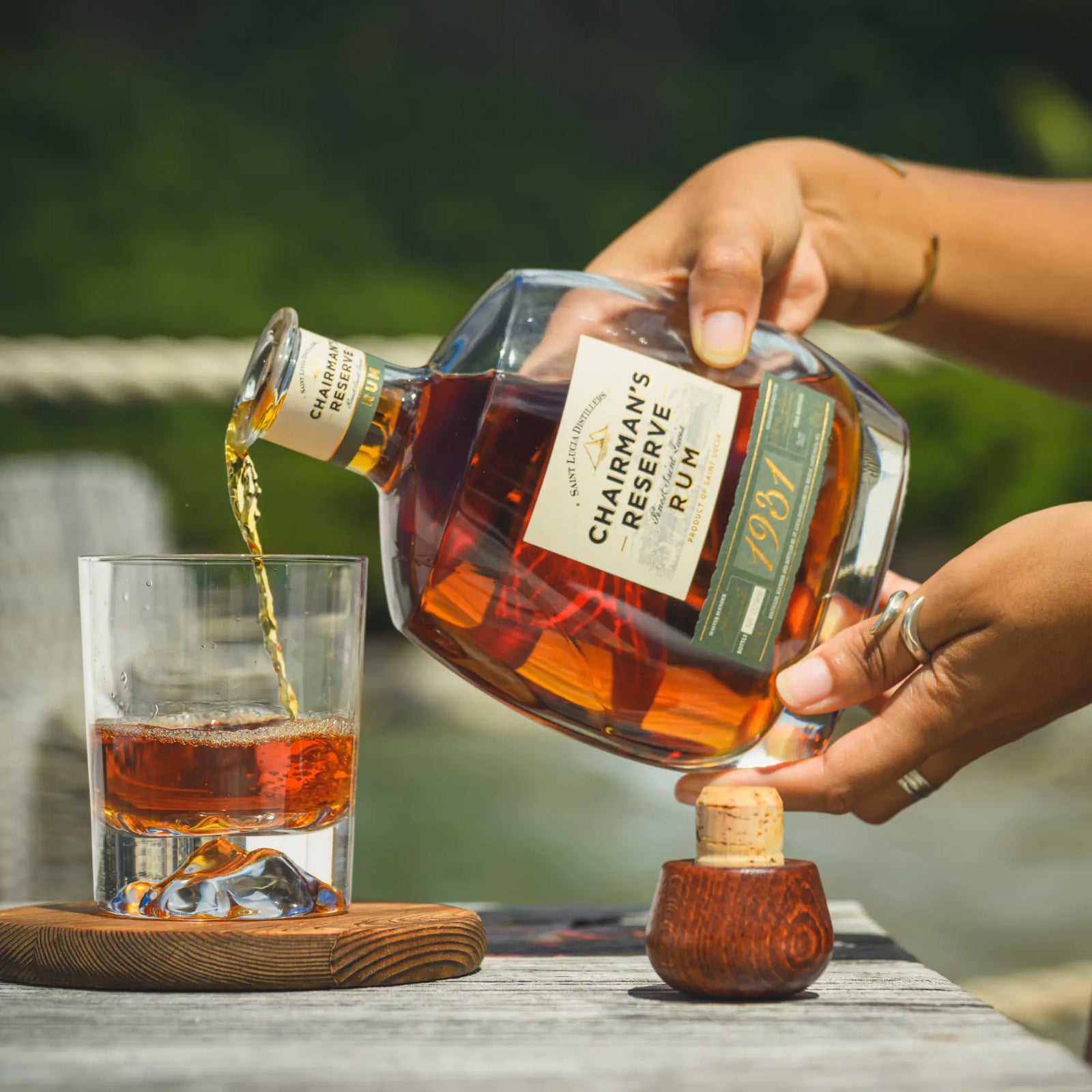 Outdoors on a wooden table, someone pours Chairman’s Reserve 1931 Rum by Chairman’s Reserve Rum over ice. The label is visible on the bottle, a cork rests nearby, and green foliage blurs softly in the background.