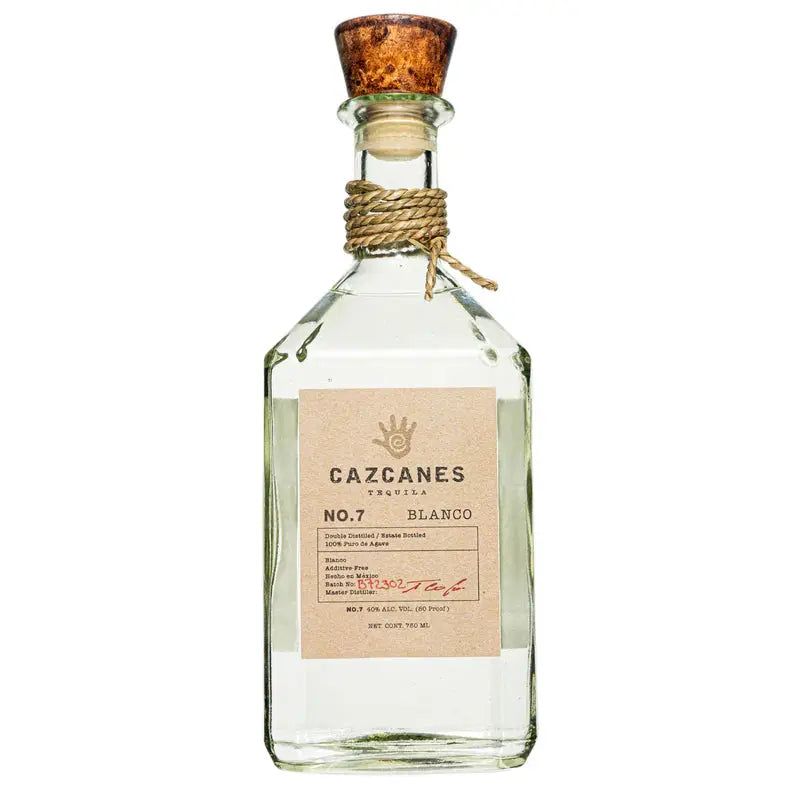 A clear glass bottle of Cazcanes No.7 Blanco Tequila by Cazcanes Tequila features a cork stopper, tan label with black and red text, and a rope around the neck. This additive-free tequila comes in a sharp-edged rectangular bottle.