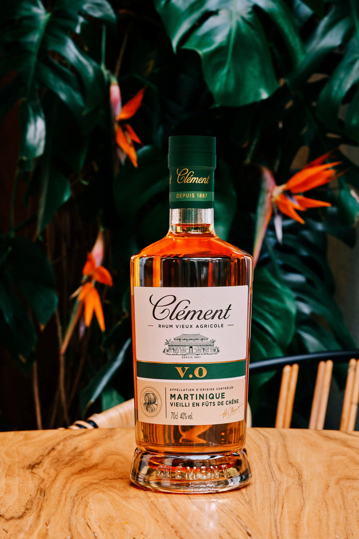 A bottle of Rhum Clément V.O by Rhum Clément rests on a wooden table, framed by a leafy green plant and bright orange bird of paradise flowers in the background.