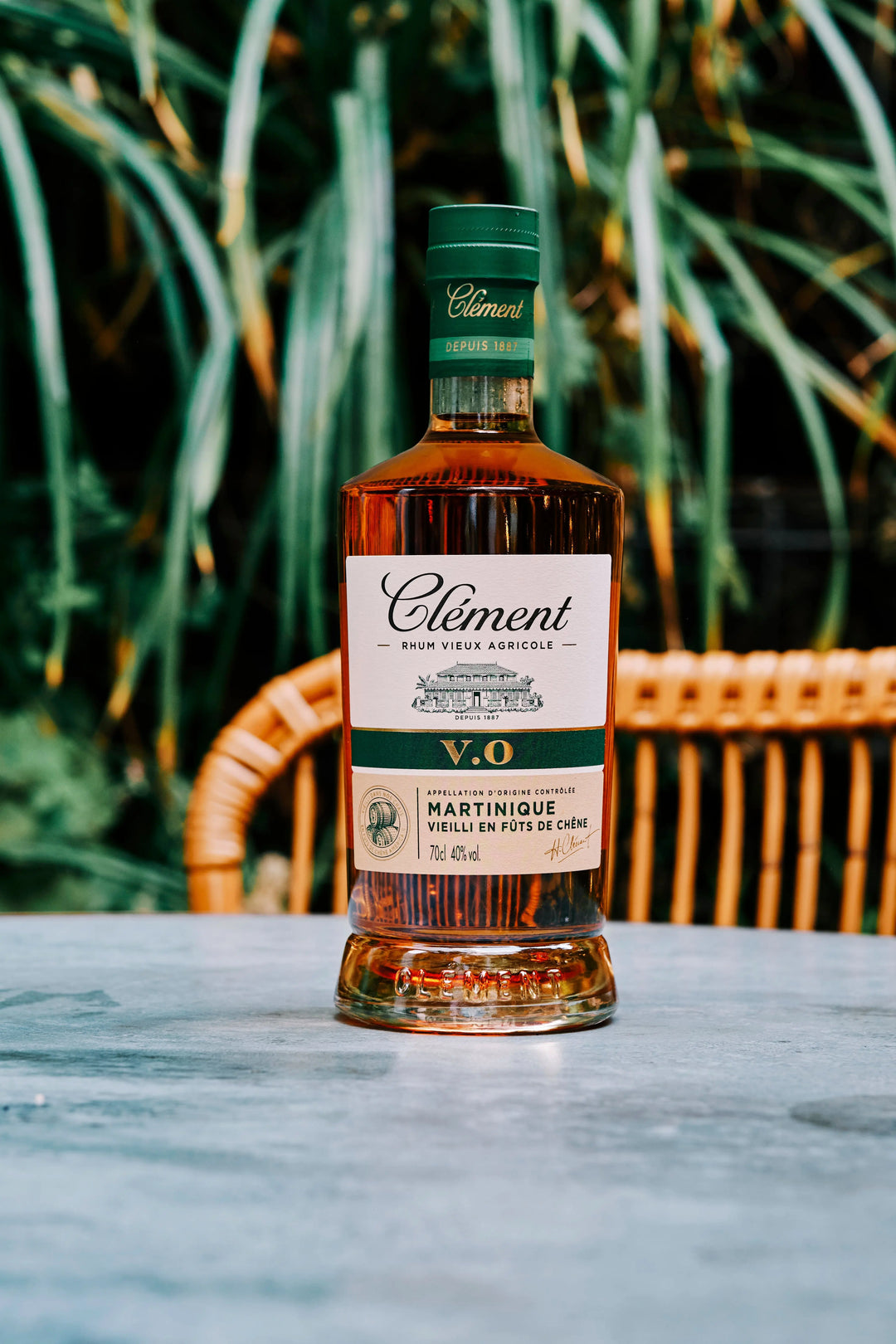 A bottle of Rhum Clément V.O, a Martinique rum made from pure sugarcane juice, rests on a marble table with a wicker chair and green plants in the background.