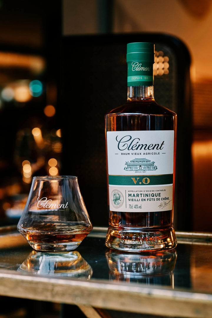 A bottle of Rhum Clement V.O by Rhum Clément, made from pure sugarcane juice, and a matching glass with the brand’s logo rest on a reflective surface under warm lighting.