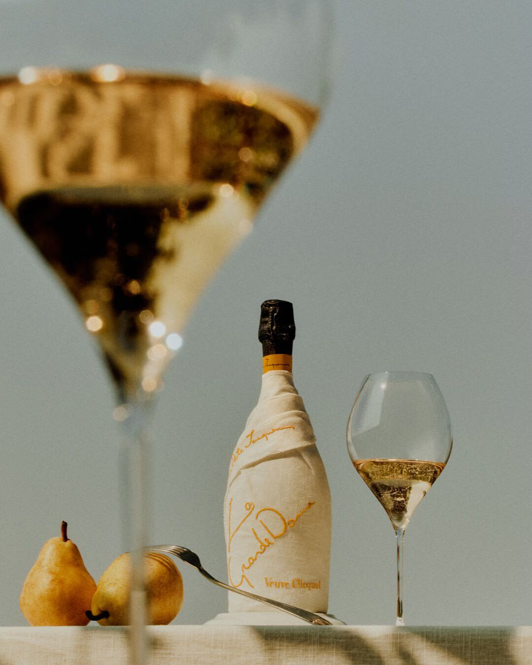 A bottle of Veuve Clicquot La Grande Dame 2018 Limited Edition by Simon Porte Jacquemus, in tissue wrap, sits with two champagne glasses and two pears on a white surface against a soft, neutral backdrop.