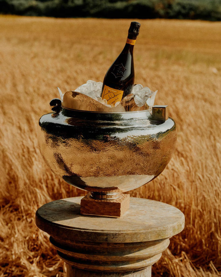 A bottle of Veuve Clicquot La Grande Dame 2018 Limited Edition by Simon Porte Jacquemus chills in a silver ice bucket on a stone pedestal amid golden wheat—an exclusive Veuve Clicquot champagne moment.