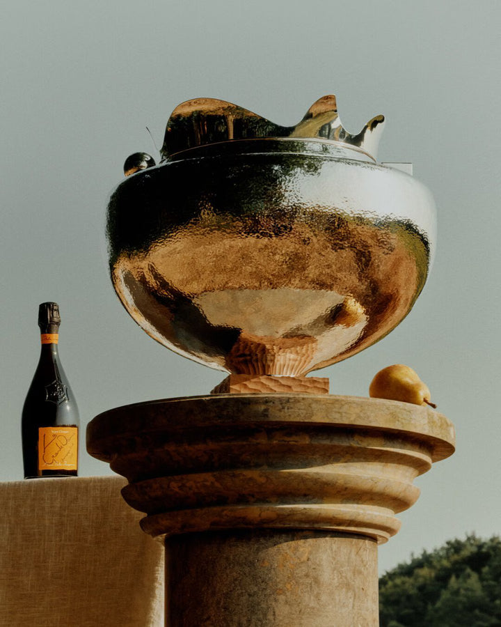 A golden bowl on a stone pedestal sits beside a lemon, with Veuve Clicquot La Grande Dame 2018 Limited Edition by Simon Porte Jacquemus in the background, all set outdoors beneath a clear sky.