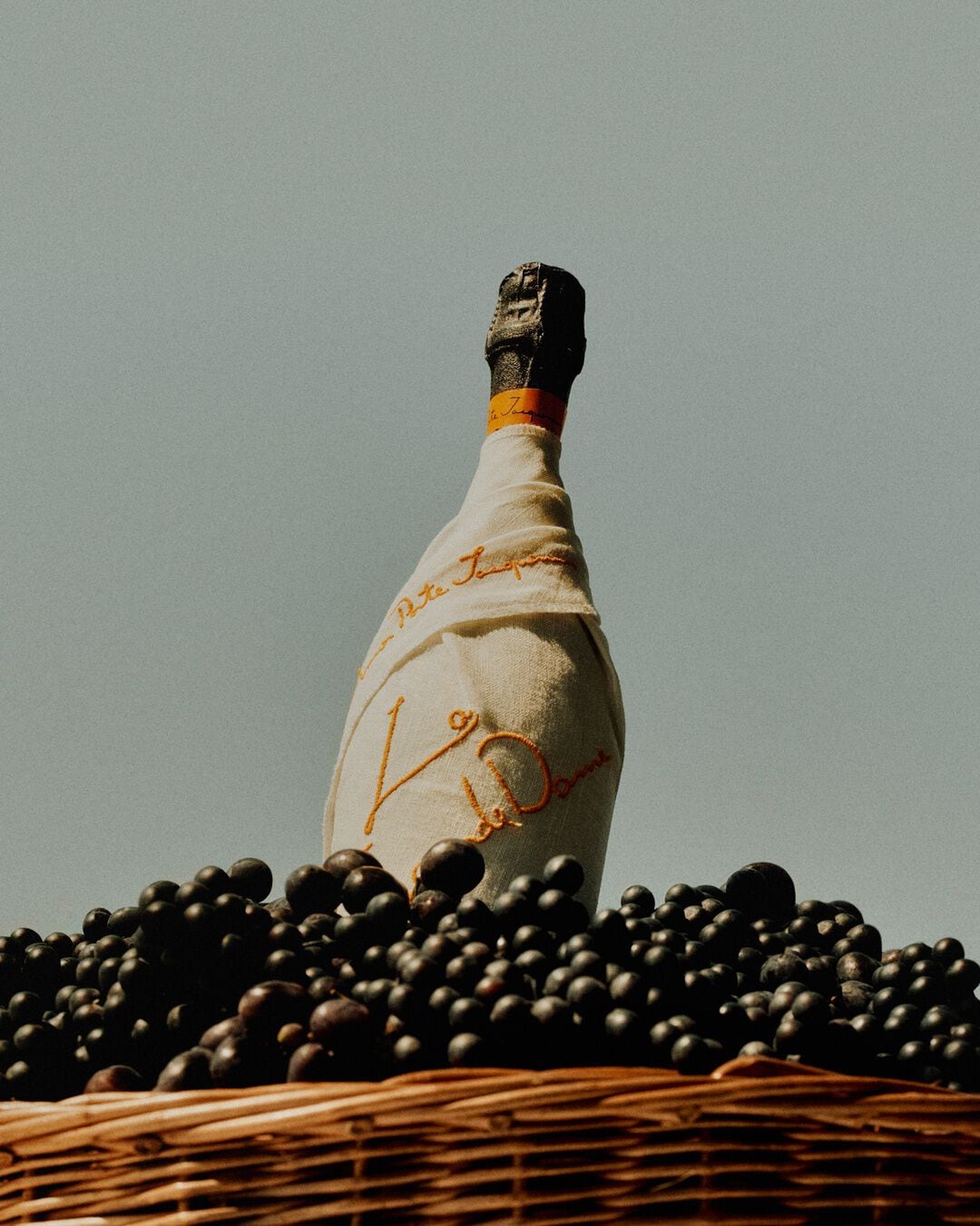 A wine bottle wrapped in cloth stands among dark grapes in a woven basket beneath a clear sky, evoking the elegance of Veuve Clicquot La Grande Dame 2018 Limited Edition by Simon Porte Jacquemus.