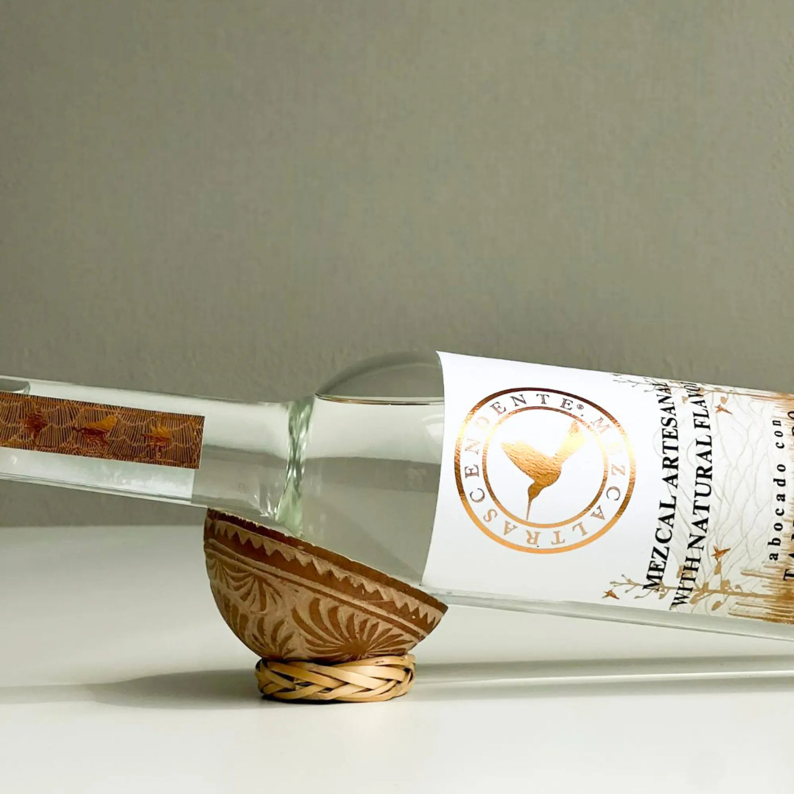 A clear bottle of Mezcal Trascendente Tamarindo 700ml with a white and gold label rests horizontally on a carved wooden stand with leaf patterns, set against a plain light background, highlighting its rich tamarind flavors.