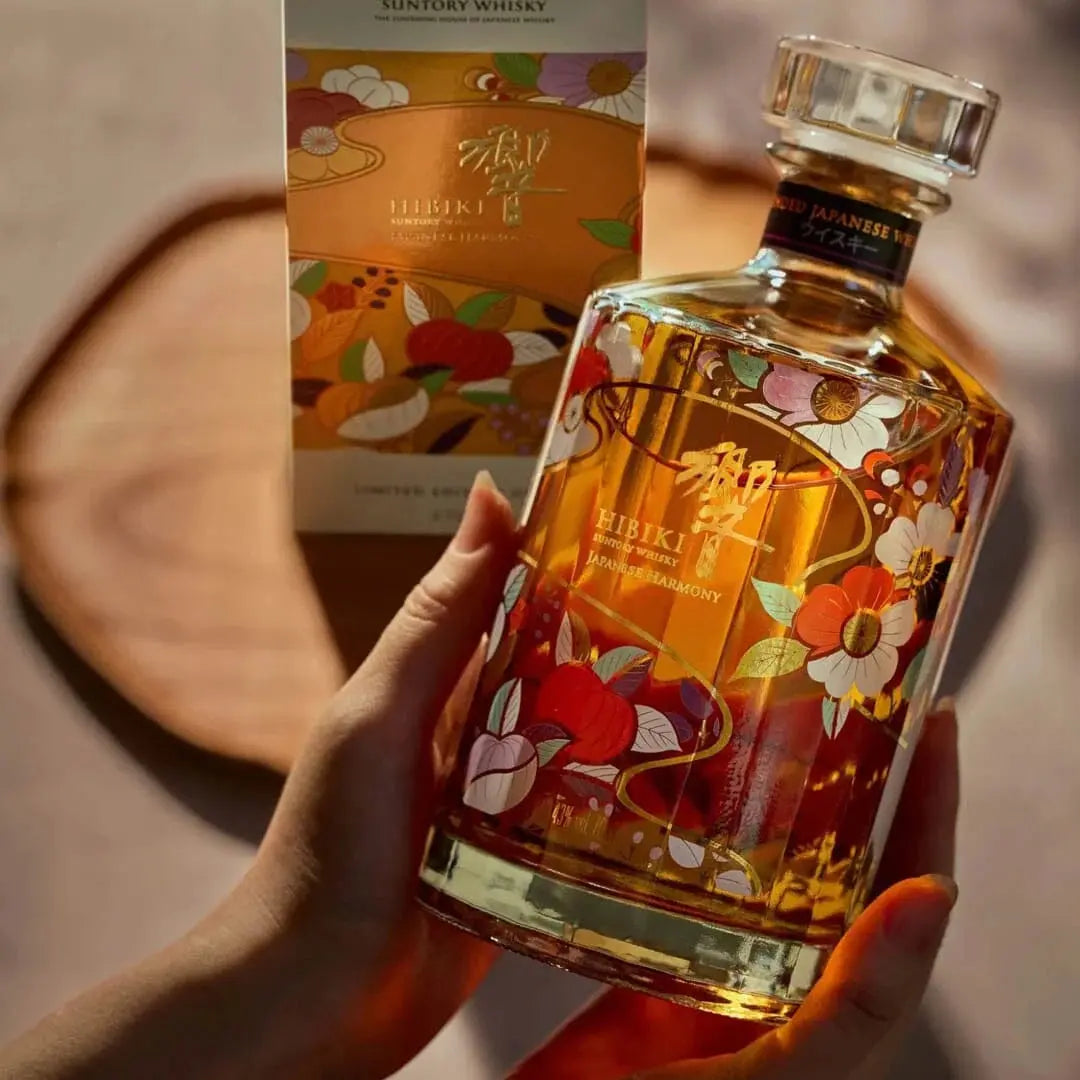 Hands holding a Hibiki Japanese Harmony Chiso 2026 Limited Edition Blended Japanese Whisky 750mL bottle decorated with colorful floral illustrations, with its matching gift box standing behind it on a wooden board.
