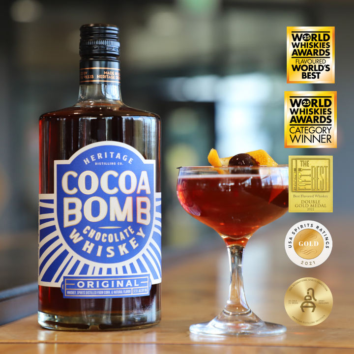 Two bottles of Heritage Distilling Cocoa Bomb Whiskey are placed next to a chocolate whiskey cocktail garnished with an orange twist and cherry, while several award medals and badges are displayed on the right.
