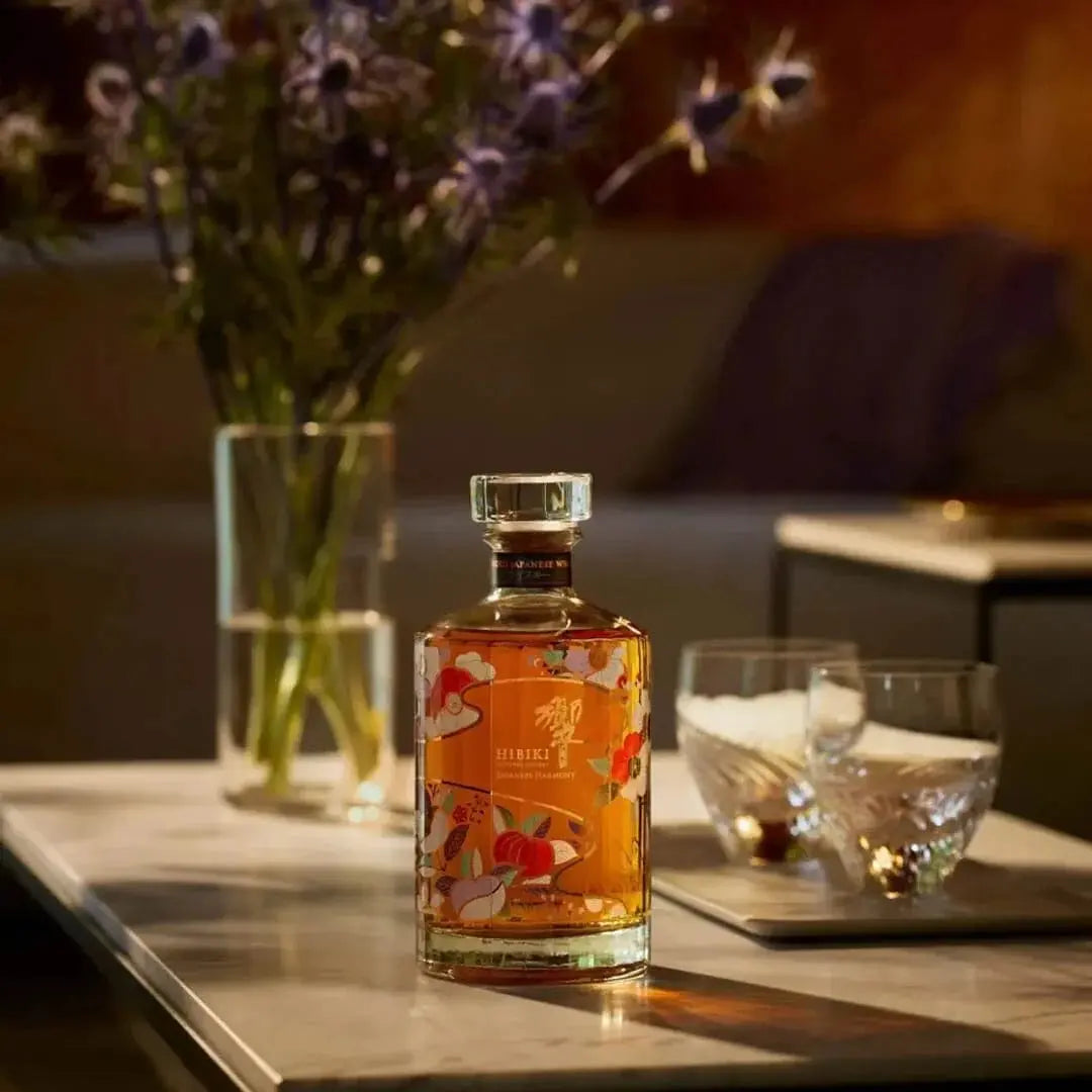 A glass bottle of Hibiki Japanese Harmony Chiso 2026 Limited Edition Blended Japanese Whisky 750mL decorated with colorful floral motifs, sitting on a table next to two empty drinking glasses and a vase of flowers.
