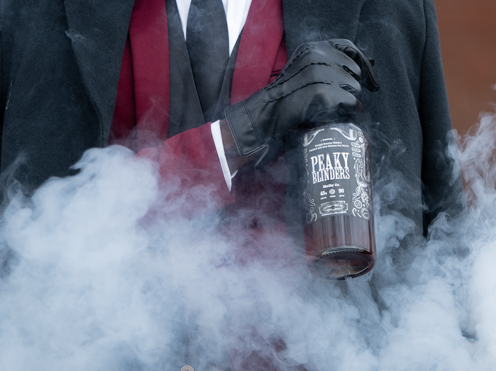 A person in a red suit, black cape, gloves, and tie holds a bottle of Peaky Blinders Irish Whiskey Shelby Company Limited Edition amid swirling mist.