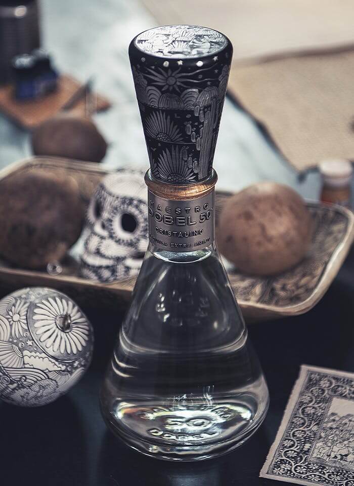 A bottle of Maestro Dobel 50 tequila with an intricately patterned black and silver cap sits on a table, surrounded by decorative spheres and objects with similar detailed black and white designs.