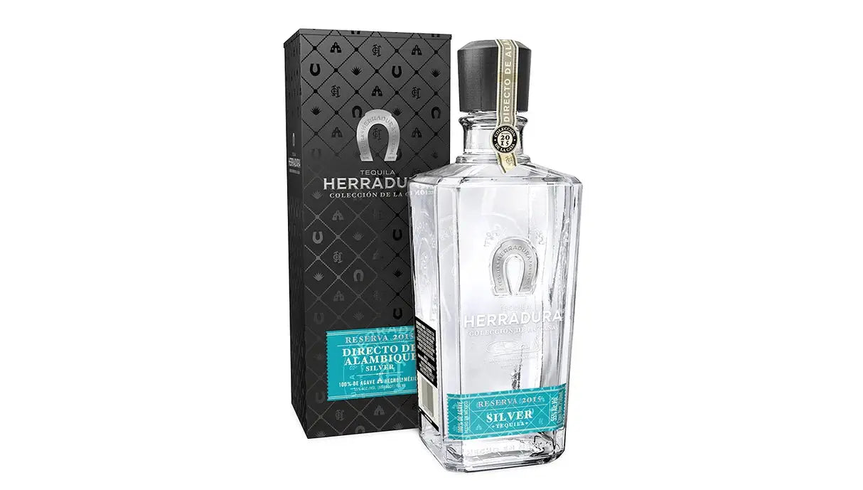 A clear bottle of Herradura Directo de Alambique stands beside its black, patterned box. Both display silver and turquoise labels with the Herradura Tequila branding and product details.