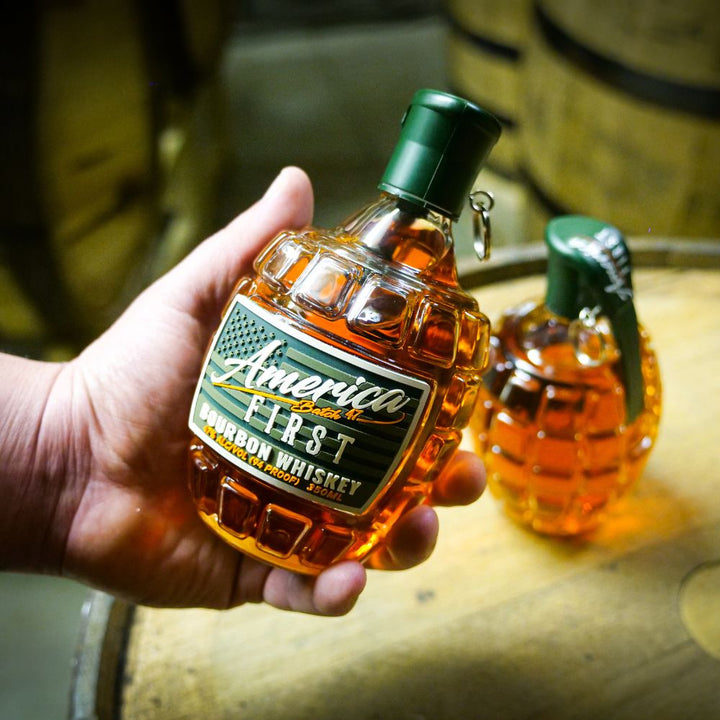 A hand holds an America First Grenade Straight Bourbon Whiskey bottle by America First, shaped like a grenade, with another in the background—ideal for patriotic gifting. Available as a 2 Pack.