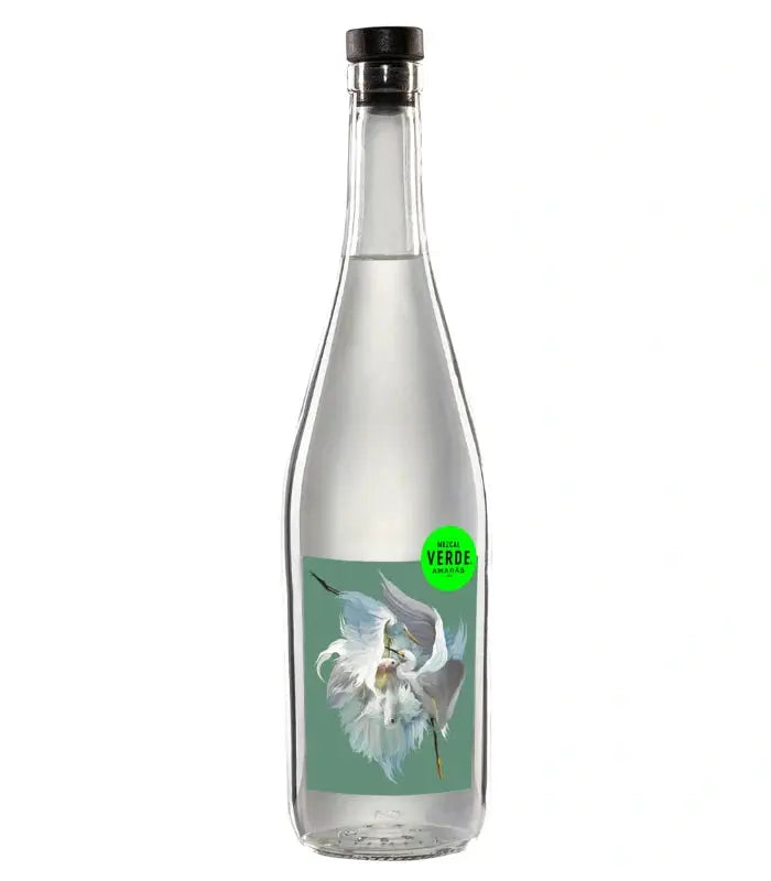 A clear glass bottle with a black cap, filled with clear liquid. The label shows a white flower on green and a small green circle reading MEZCAL VERDE. Product: Amaras Mezcal Verde Artist Series 2024 by Amaras.