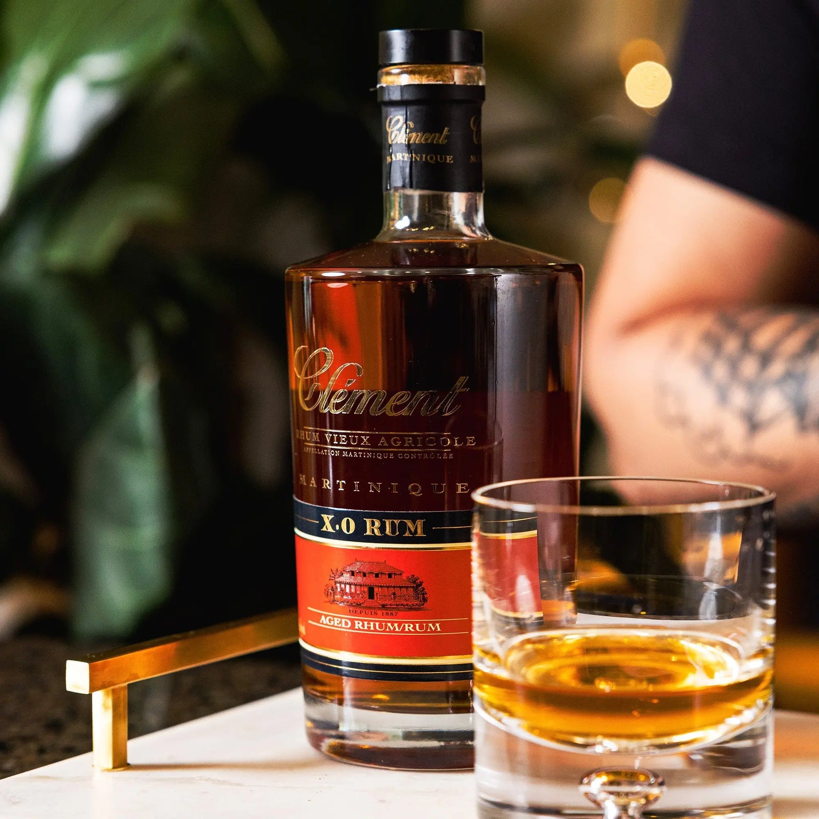 A bottle of Rhum Clément X.O. Vieux Rhum Agricole from Rhum Clément stands beside a glass on the table, while a tattooed arm in the background enhances the scene’s warm, inviting vibe.