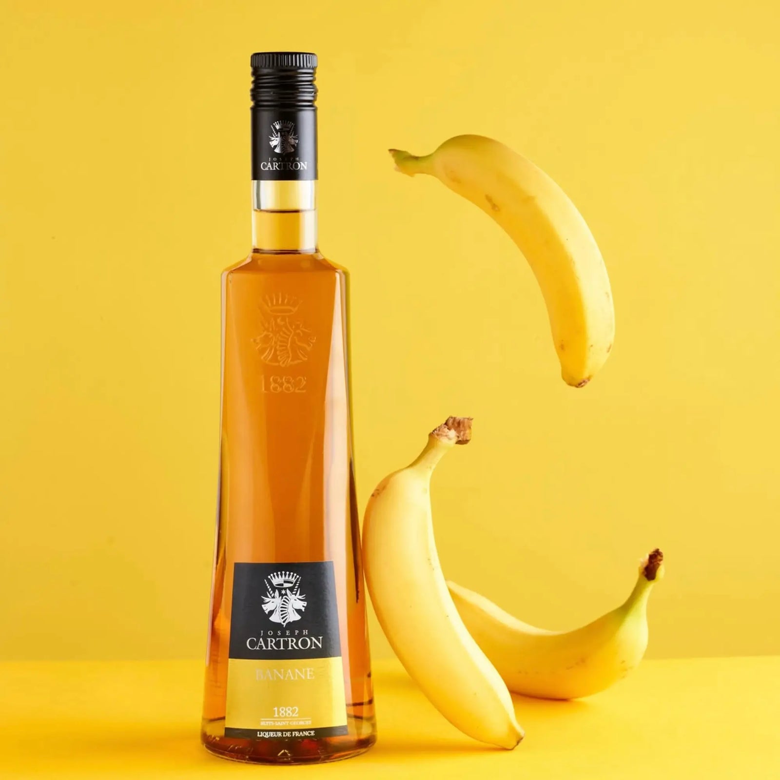 A 700 ml bottle of Cartron Banane (Banana) Liqueur stands on a yellow surface with three Ecuadorian bananas—two resting and one floating—set against a solid yellow background, capturing the brand's distinctive banana flavor.