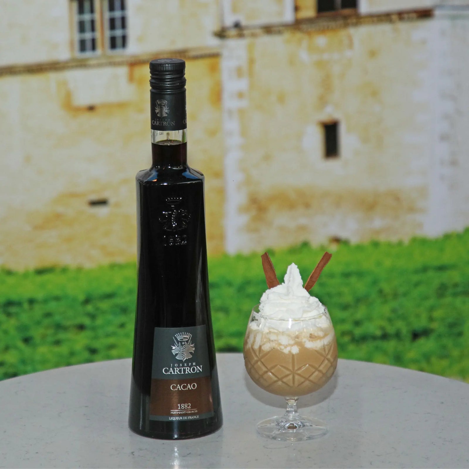 A bottle of Cartron Cacao Brun Liqueur 700 sits beside a creamy, whipped-topped drink with cinnamon sticks on marble—crafted from Ivory Coast cocoa beans, with hints of Cognac from the Cartron brand filling the air.