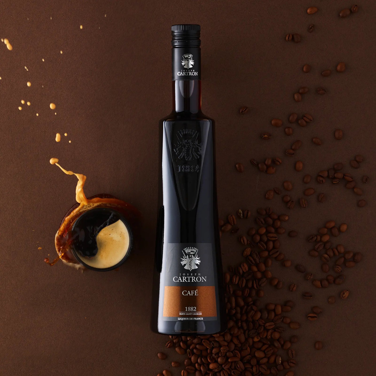 A bottle of Cartron Café (Coffee) Liqueur 700 ml sits on a brown surface beside a tipped espresso cup spilling coffee, surrounded by scattered Costa Rica coffee beans.
