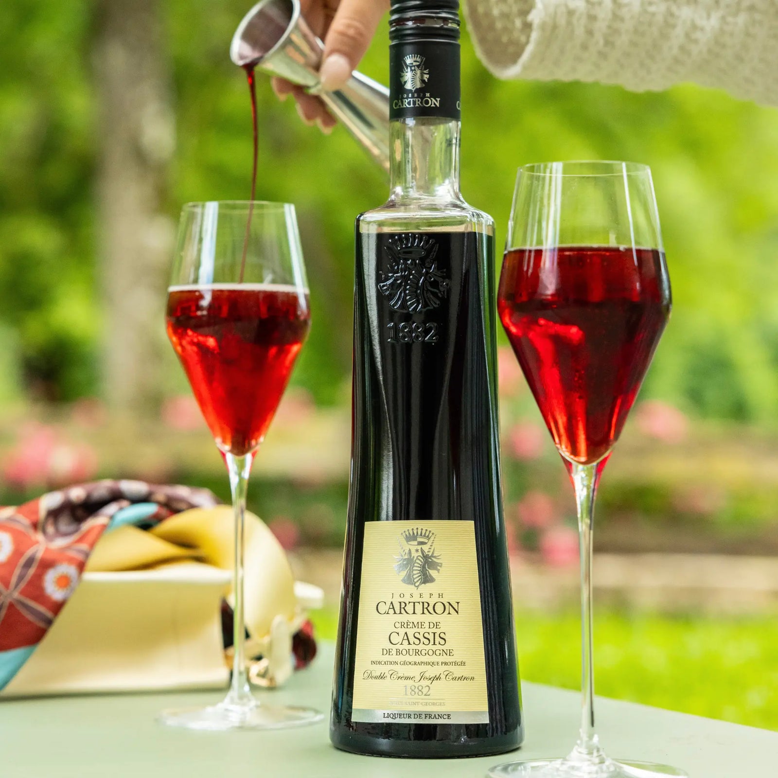 Someone pours deep red Cartron Crème de Cassis de Bourgogne (Blackcurrant) Liqueur 700 ml into a glass beside its tall bottle and another filled glass, all arranged outdoors with greenery and flowers softly blurred behind.