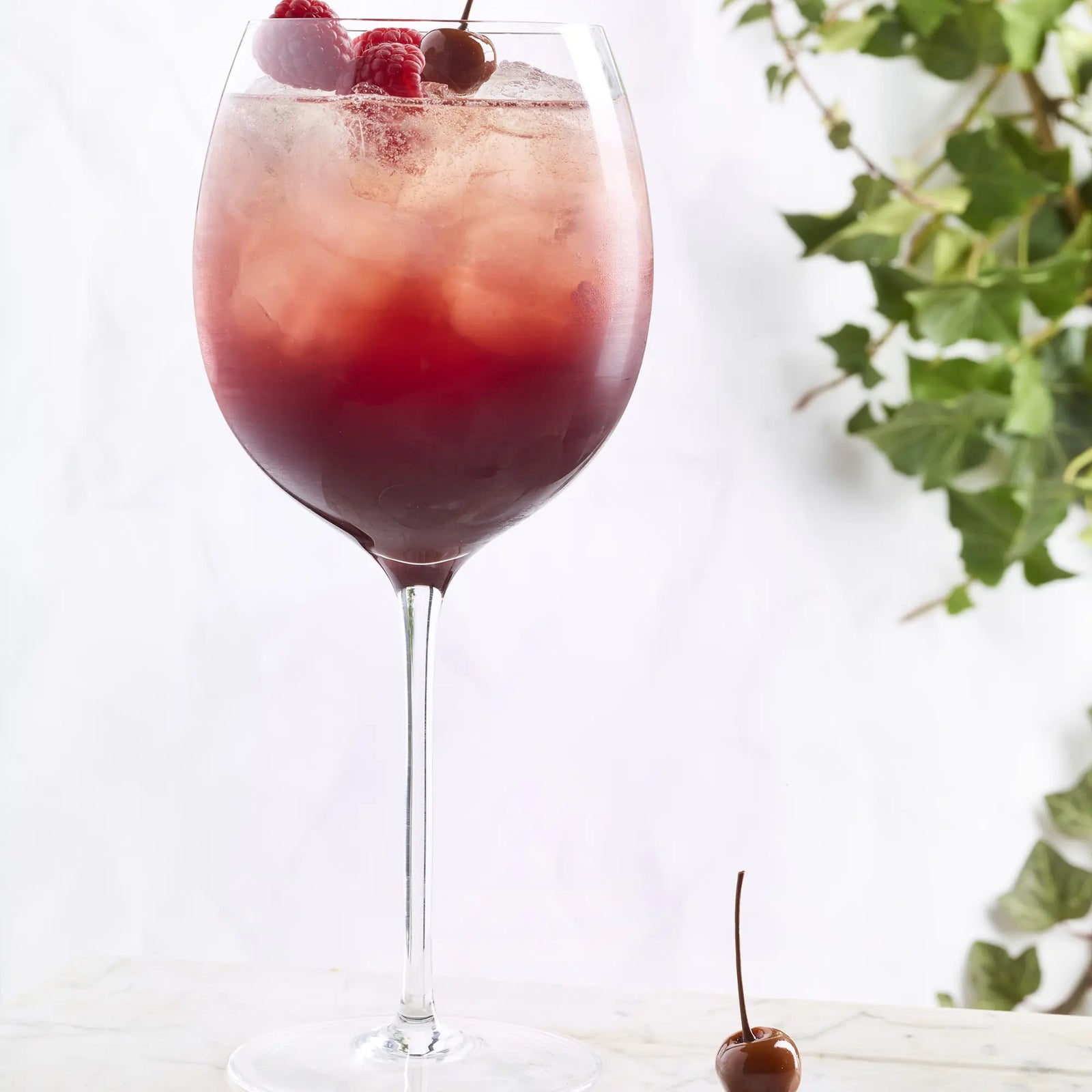 A large wine glass holds a red iced drink made with Cartron Crème de Cerise (Cherry) Liqueur 700ml, garnished with raspberries and local cherries. A cherry sits nearby, with green ivy and hints of Burgundy fruit in the background.