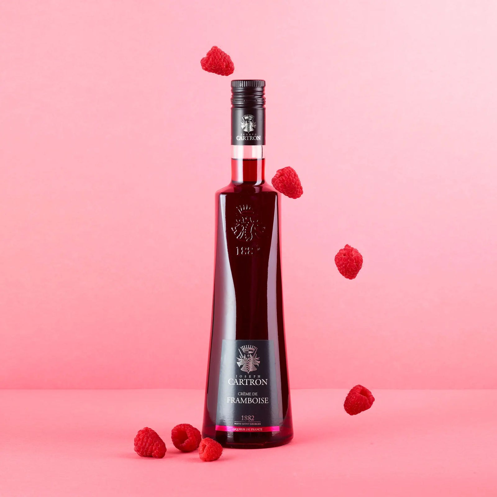 A tall, elegant bottle of Cartron Crème de Framboise (Raspberry) Liqueur 700 ml stands upright against a pink background, surrounded by fresh raspberries, some resting on the surface and others appearing to float in the air.