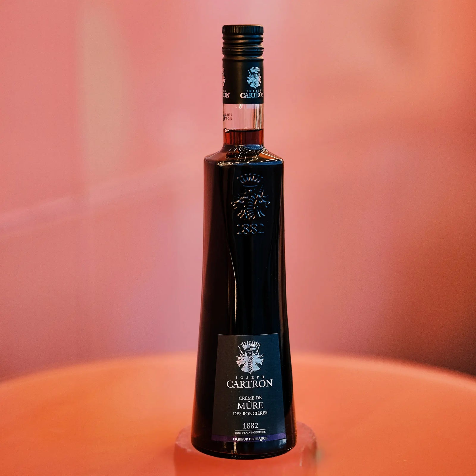 A sleek bottle of Cartron Crème de Mûre (Blackberry) Liqueur 700 ml by Cartron stands on a glossy pink surface against a soft pink background.