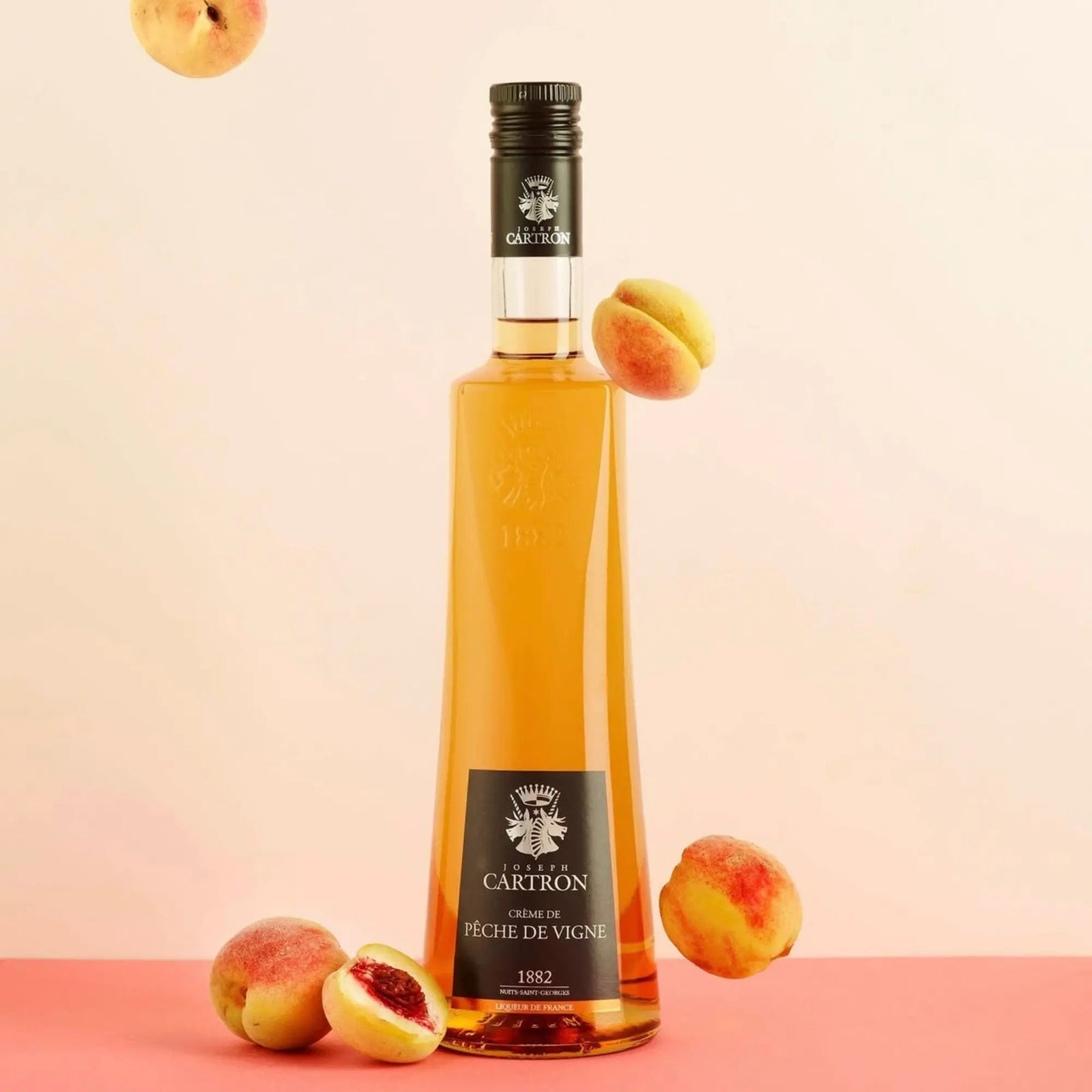 A 700 ml bottle of Cartron Crème de Pêche de Vigne Peach Liqueur stands upright, surrounded by whole and halved Bourgogne peaches, set against a peach and pink background.