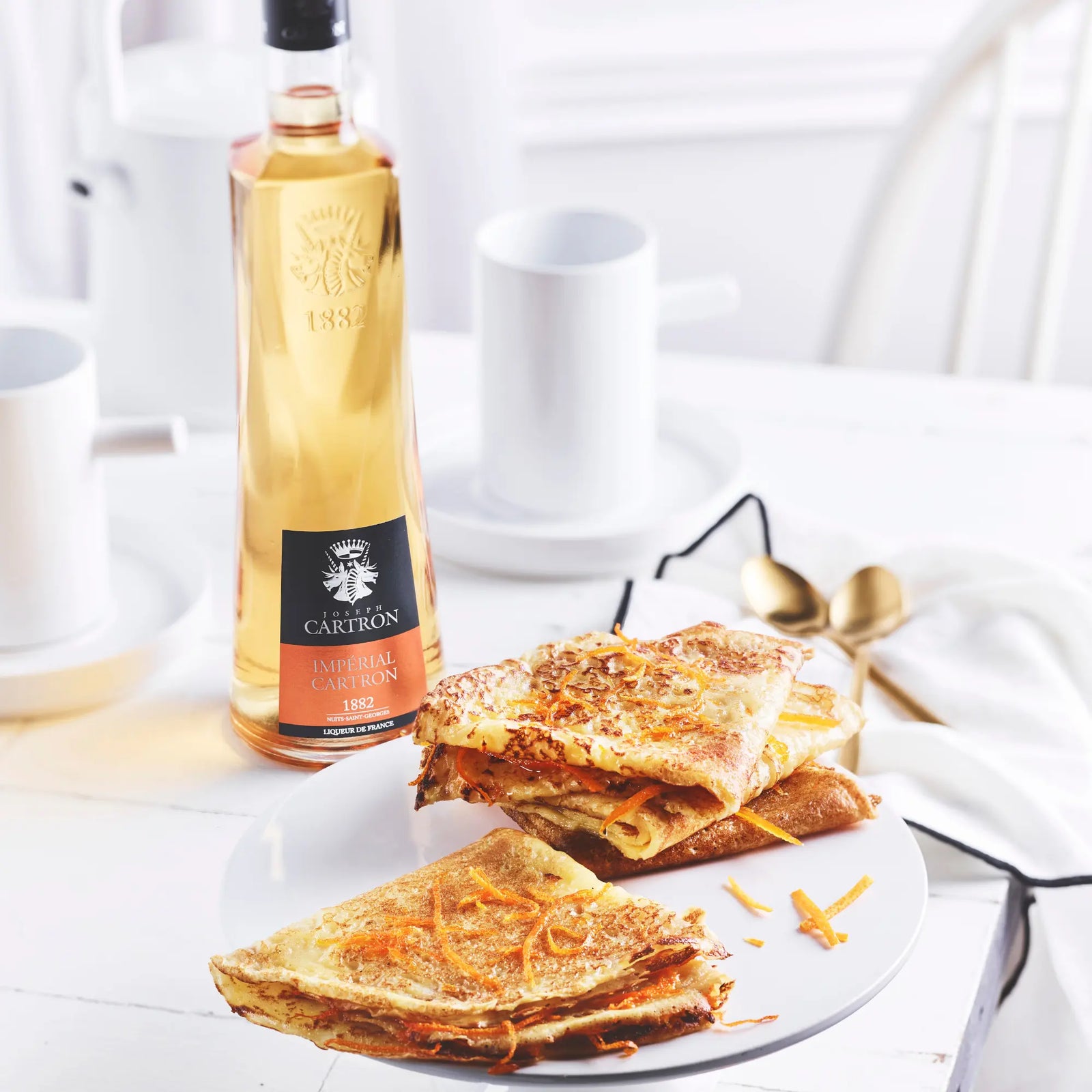 Folded crepes with orange zest sit on a white table beside a bottle of Cartron Impérial Cartron Triple Orange Liqueur 700 ml, with white cups, saucers, and golden spoons in the background for an elegant breakfast scene.