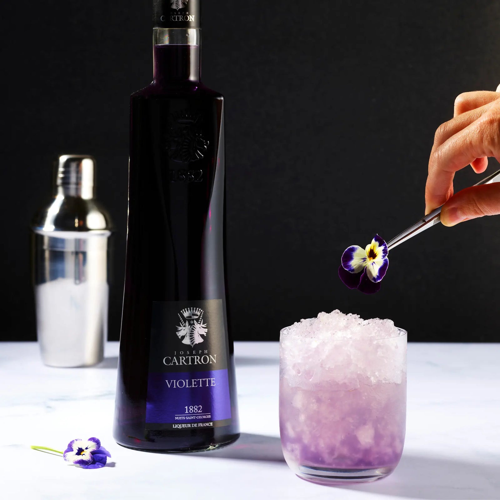 A bottle of Cartron Violette Liqueur 700 ml stands beside a glass with crushed ice and a purple cocktail. A hand adds a violet leaf garnish, emphasizing the floral aroma. The Cartron brand shines as a shaker appears in the background.
