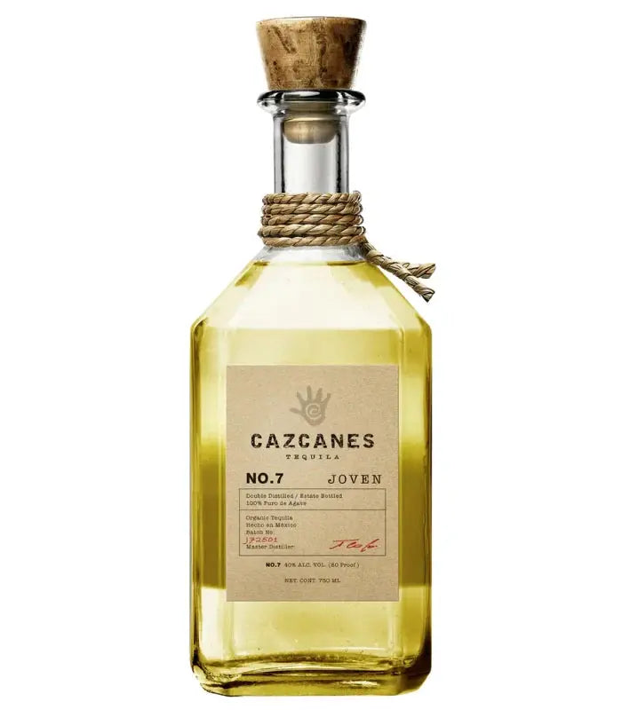 A clear glass bottle of Cazcanes No. 7 Tequila Joven with a cork stopper, tan label, and rope around the neck displays pale yellow tequila inside.