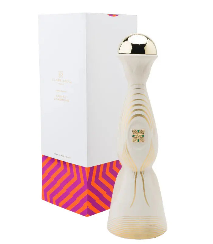 A stunning Clase Azul Tequila Joven Spirit Of Champions Limited Edition bottle in white and gold ceramic with a rounded gold cap is displayed before its box, featuring bold pink and orange geometric designs at the base.