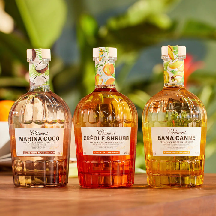 Three glass bottles of Rhum Clément French Caribbean liqueurs—Mahina Coco, Créole Shrubb, and Bana Canne Liqueur—are displayed on a wooden surface with leafy greenery softly blurred in the background.