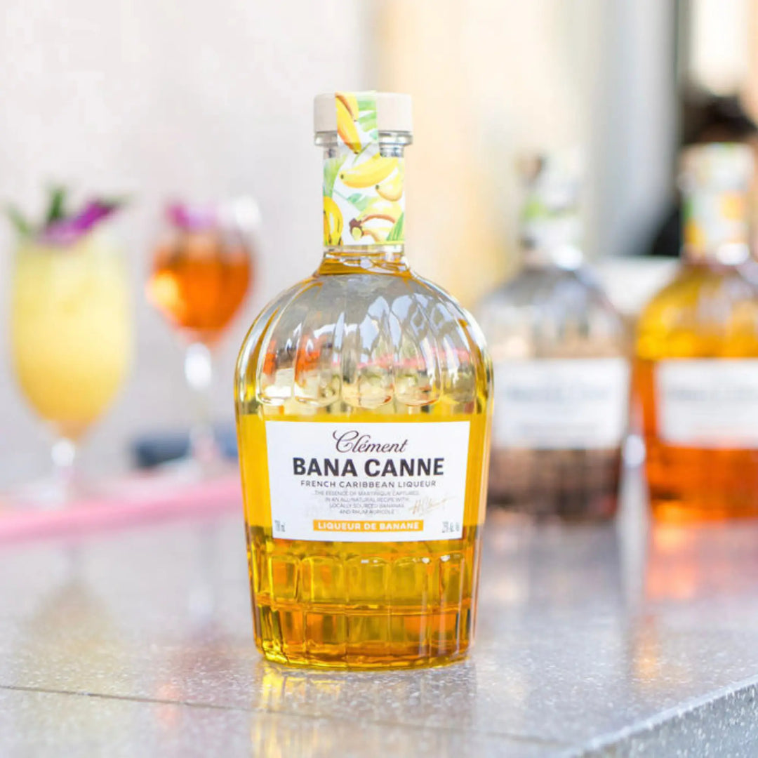 A bottle of Rhum Clément Bana Canne Liqueur from Rhum Clément rests on a table, its label adorned with gold accents and bananas, while cocktails and other bottles create a blurred backdrop. The design hints at the liqueur&