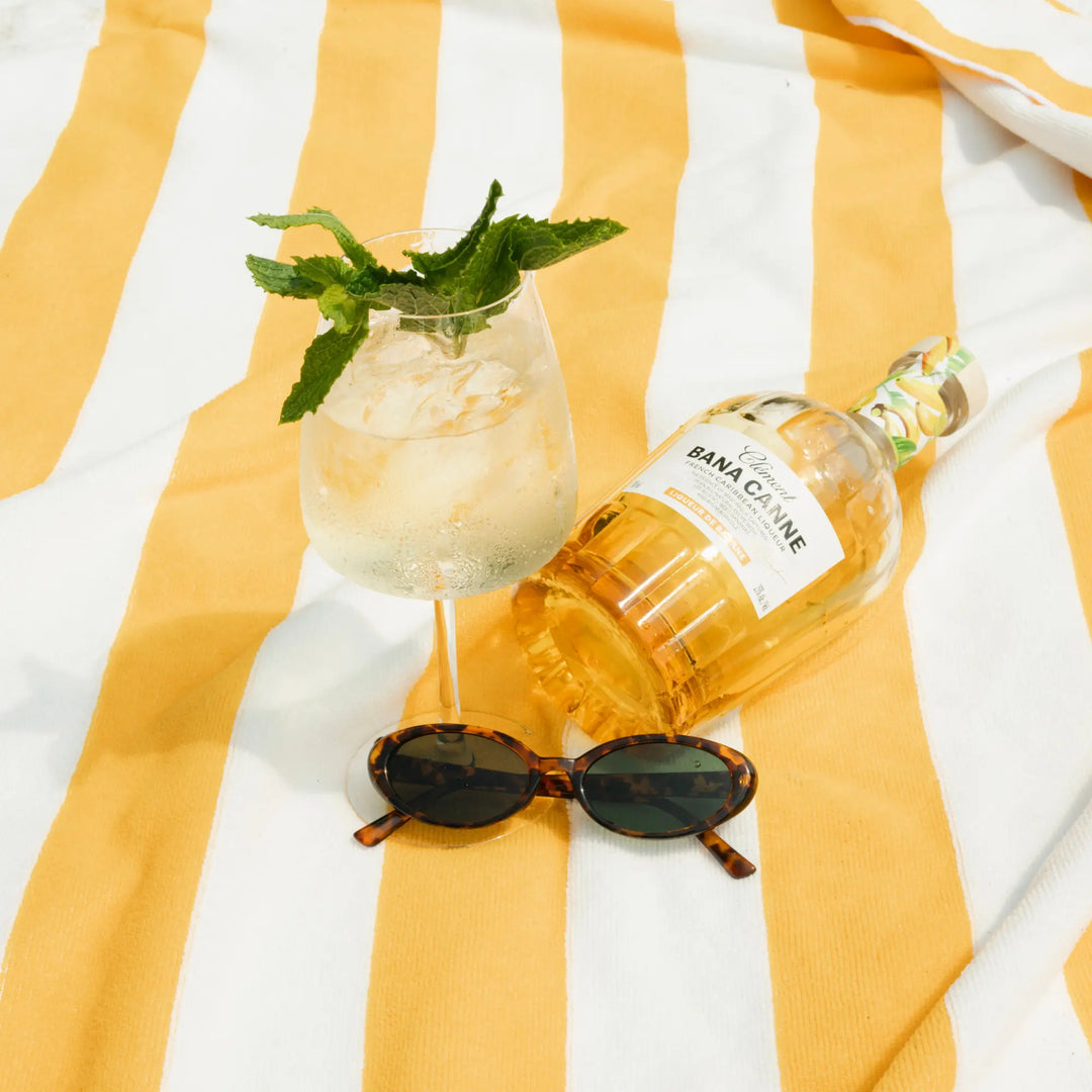 A glass of iced cocktail garnished with mint, a bottle of Rhum Clément Bana Canne Liqueur, and tortoiseshell sunglasses sit on a yellow and white striped beach towel.