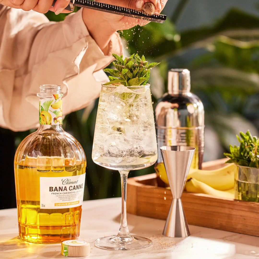 A hand grates zest over a minty cocktail in a wine glass. Nearby are bananas, fresh mint, and a bottle of Rhum Clément Bana Canne Liqueur by Rhum Clément. The bright scene highlights the natural banana essence.