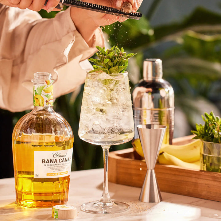 A hand grates zest over a minty cocktail in a wine glass. Nearby are bananas, fresh mint, and a bottle of Rhum Clément Bana Canne Liqueur by Rhum Clément. The bright scene highlights the natural banana essence.