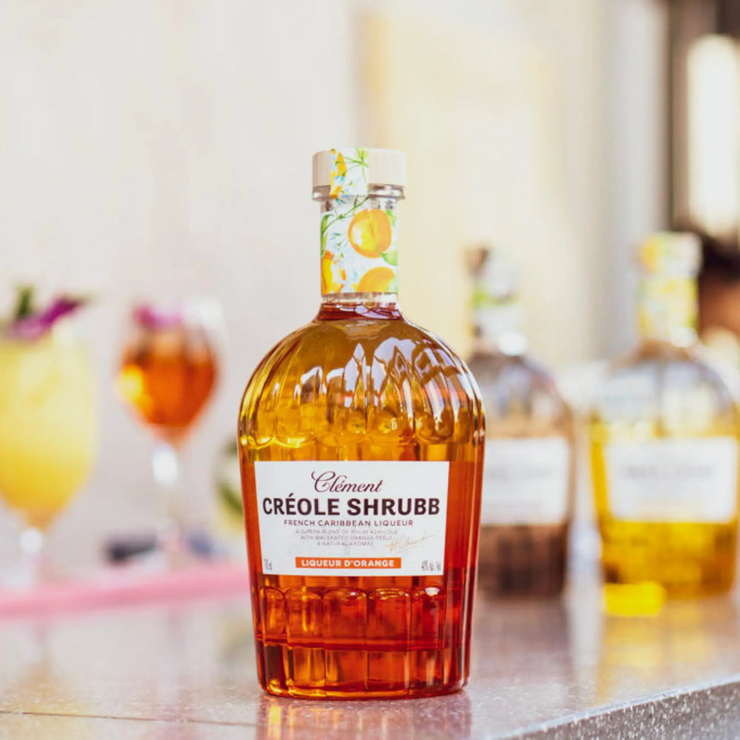 A bottle of Rhum Clément Créole Shrubb Orange Liqueur by Rhum Clément sits on a reflective surface, with two blurred bottles and vibrant cocktails in the background, creating a lively, summery vibe.