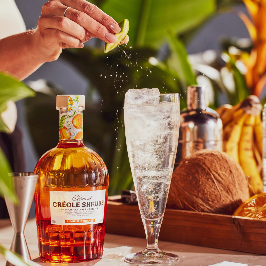 A hand squeezes a lime wedge over a tall glass of ice beside a bottle of Rhum Clément Créole Shrubb Orange Liqueur. A metal shaker, coconut, bananas, and lush greenery create a vibrant tropical atmosphere in the background.