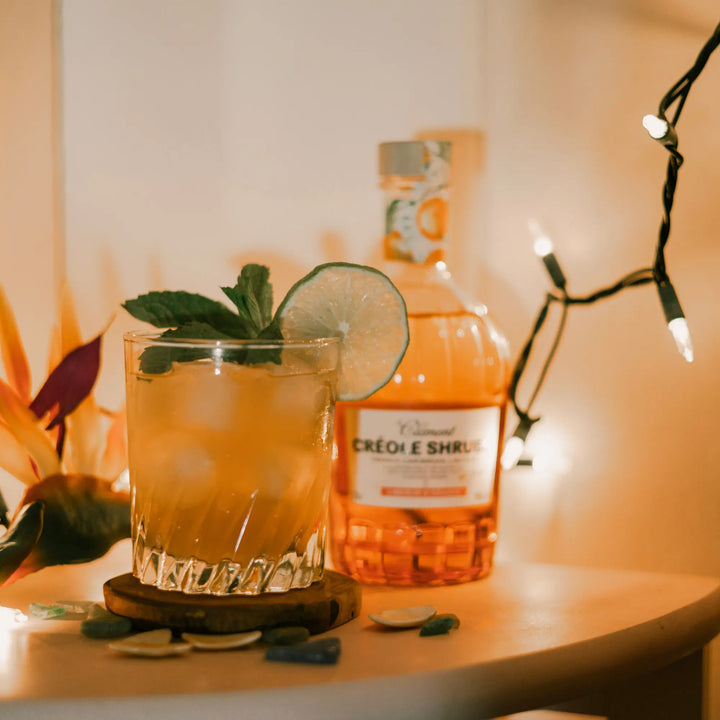 A glass of iced cocktail with lime and mint sits on a coaster beside a bottle of Rhum Clément Créole Shrubb Orange Liqueur, surrounded by flowers, colorful stones, and string lights.