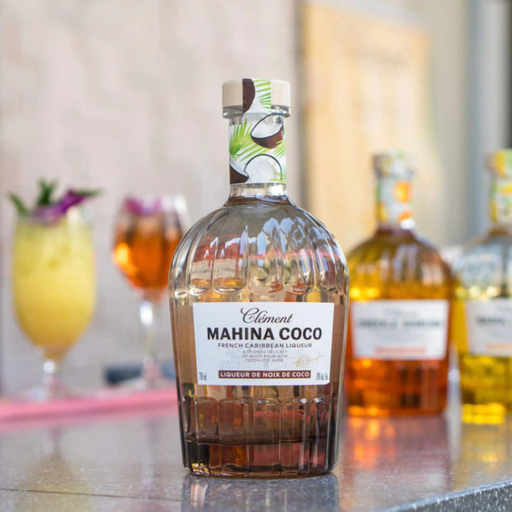 A bottle of Rhum Clément Mahina Coco | Coconut Liqueur from Rhum Clément rests on a counter, its label featuring coconuts and highlighting the blend of natural coconut liqueur and sugarcane syrup, with tropical cocktails in the background.