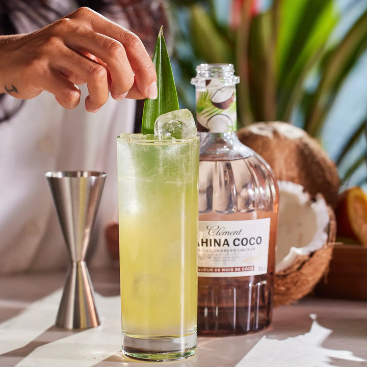 A hand adds a leaf garnish to a tall glass of yellow-green cocktail with ice. Behind it sits a bottle of Rhum Clément Mahina Coco | Coconut Liqueur from Rhum Clément, a jigger, and half a coconut on a sunlit table.