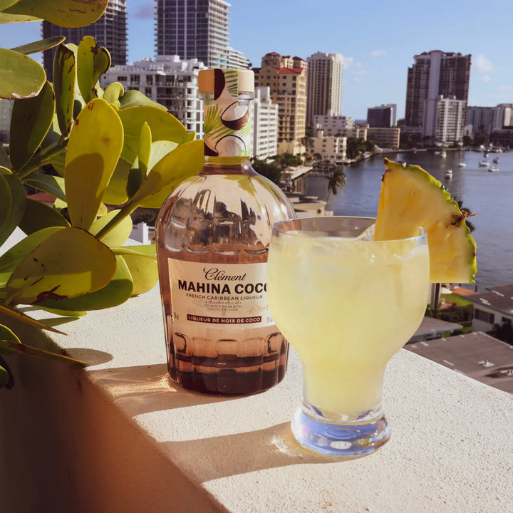 A bottle of Rhum Clément Mahina Coco Coconut Liqueur and a tropical cocktail with pineapple garnish rest on a balcony ledge, overlooking city buildings, water, and blue sky in the background.