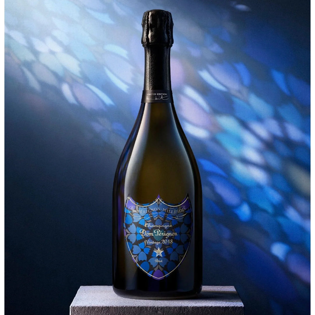 Dom Pérignon Vintage 2018 Damien Hirst Limited Edition Brut Champagne bottle with a dark glass body and a label featuring a blue and black floral motif, standing on a stone pedestal.