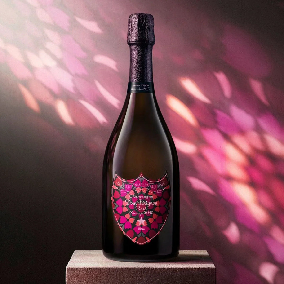 A bottle of Dom Pérignon Rosé Vintage 2010 Damien Hirst Limited Edition Champagne with a decorative, multi-colored floral label stands on a square stone pedestal against a pink and purple patterned background.