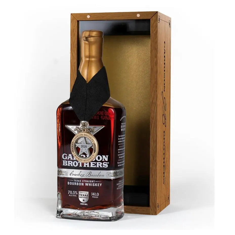 A bottle of Garrison Brothers 2026 Cowboy Bourbon Texas Straight Bourbon Whiskey, proudly bearing a medal around its neck, is showcased beside an open wooden box featuring a gold interior.