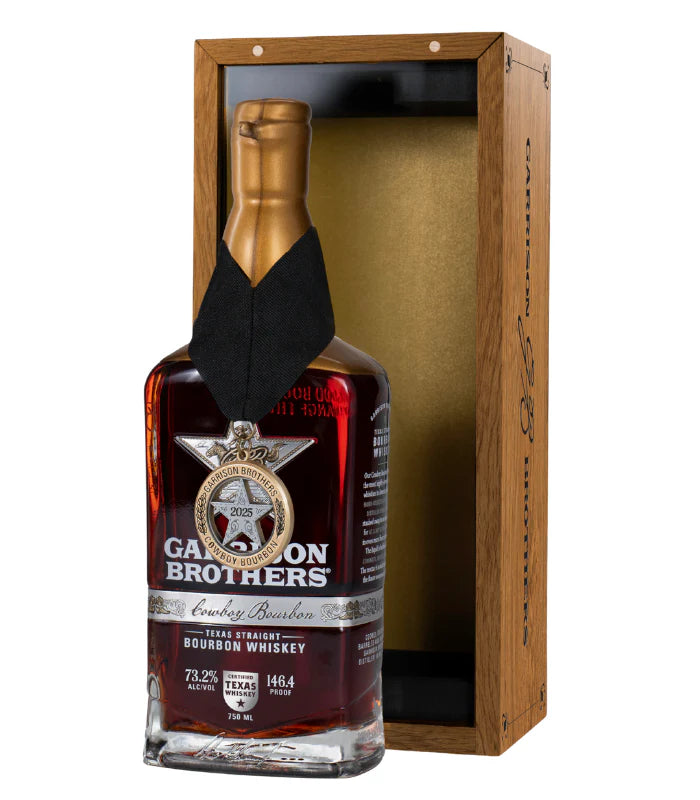 A bottle of Garrison Brothers Cowboy Bourbon 2025 Limited Edition 146.4 Proof with a gold wax seal and medallion sits before an open wooden box lined with a gold interior.