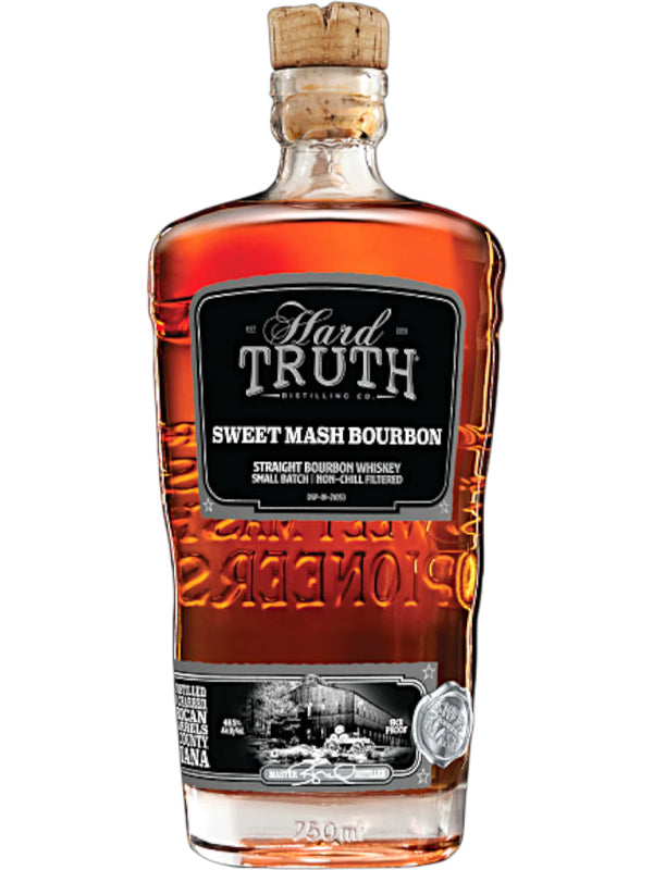 A clear glass bottle of Hard Truth Sweet Mash Bourbon Whiskey features a black label and wooden cork, displaying the Hard Truth brand name and small batch bourbon details with amber liquid visible inside.