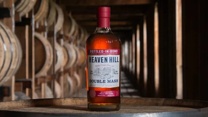 A bottle of Heaven Hill Double Mash Bottled-in-Bond Bourbon (Pre-Order) rests on a barrel inside a dimly lit rickhouse, with rows of wooden barrels stacked in the background.