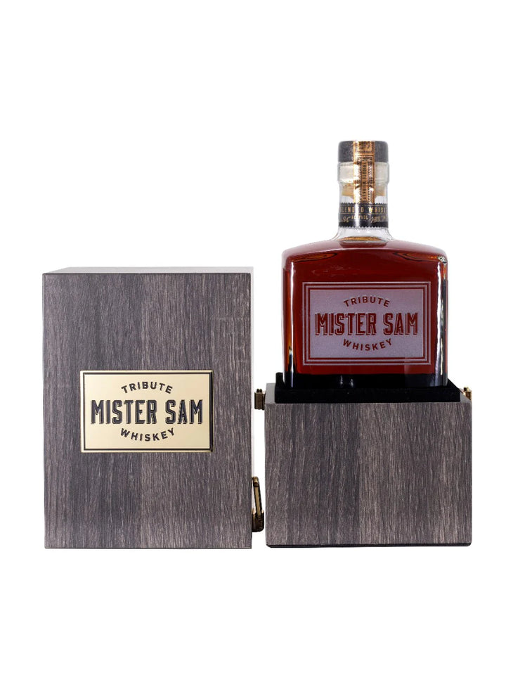 A bottle of Sazerac Mister Sam Tribute Whiskey Second Edition, a cask strength blend, comes in a wooden box with a gold label. The open box displays the bottle featuring its bold red and gold “Mister Sam Tribute Whiskey” label.