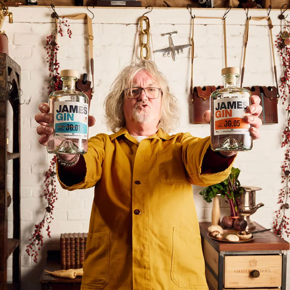 A gray-haired man in glasses and a yellow jacket stands indoors, holding two bottles of James Gin The Internationalist W/ Souvenir Gift Key Chain by James Gin US. Tools, hanging herbs, and vintage decor are visible in the background.